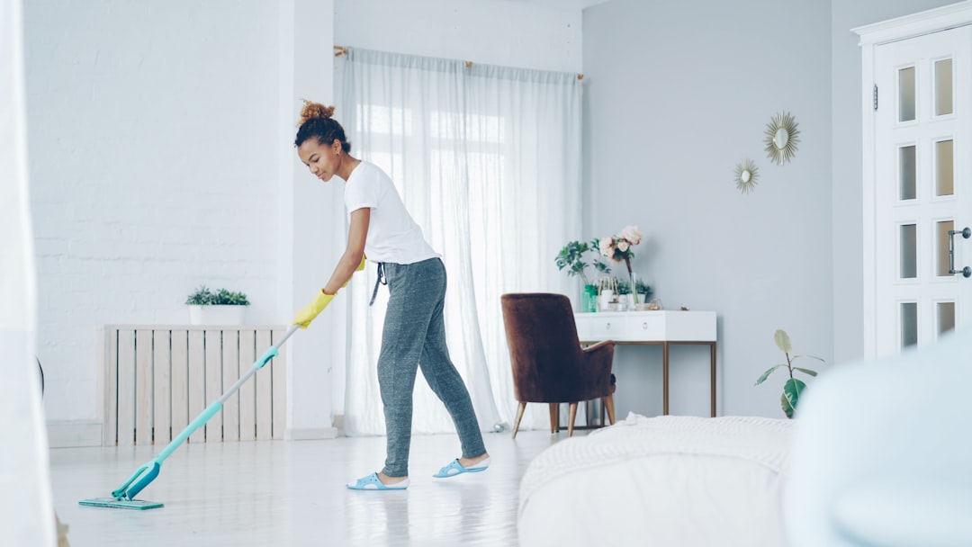 Pretty African American girl is mopping floor at home with flat plastic mop wearing gloves and wear-at-home clothing. Modern interiors, cleanness and young people concept.